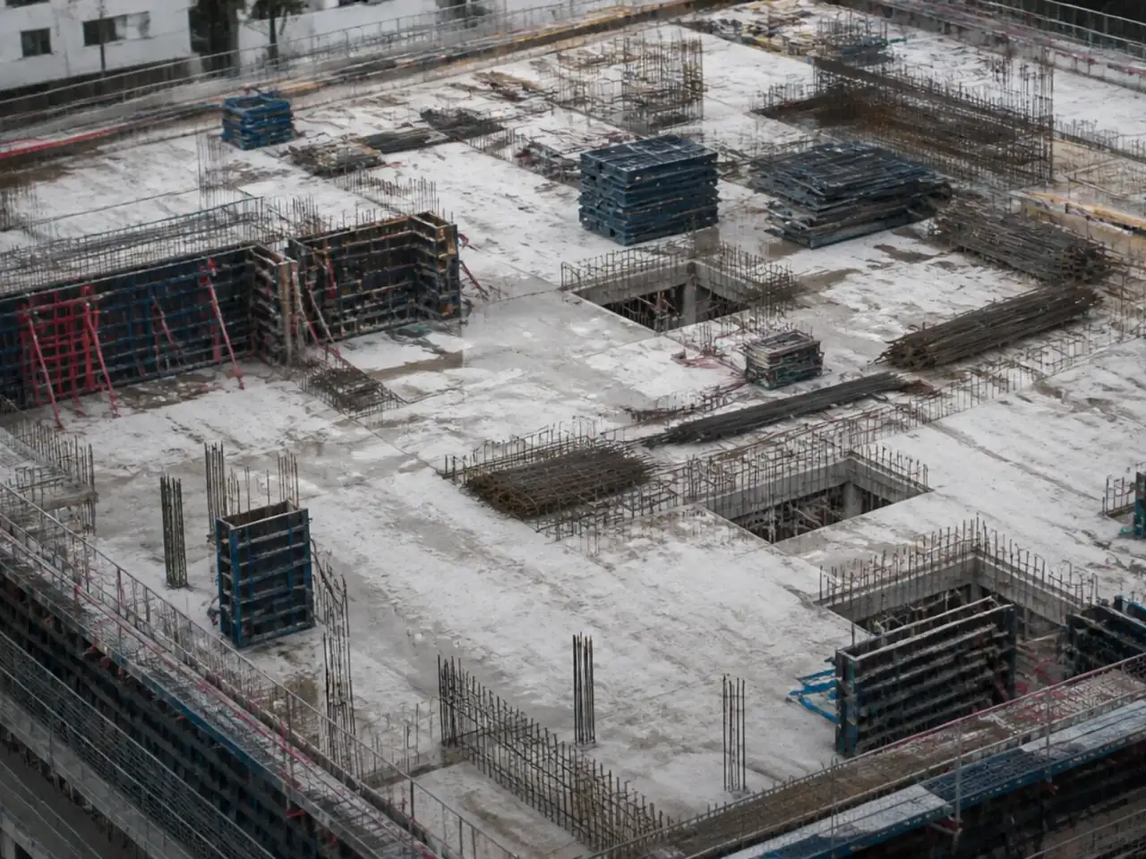 Vue plongeante lointaine sur une dalle de chantier en cours de coffrage, ouvriers réduits à des silhouettes non identifiables
